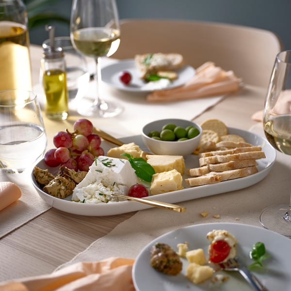 Cheese platter on large white plate with smaller plates and wine glasses, surrounded by grapes, olives, and bread. Elegant table setting.