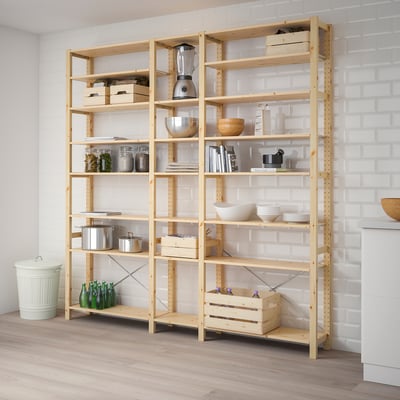 A wooden IVAR storage unit filled with kitchen items, sized similarly to a person and set against a white brick wall.