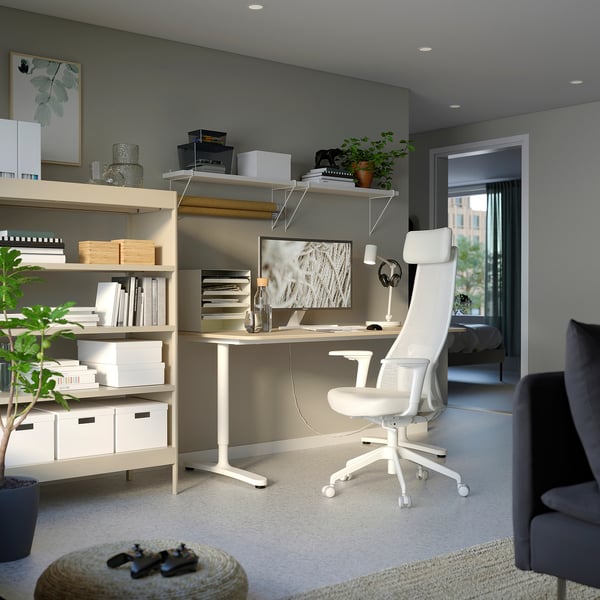 Modern home office: white ergonomic chair, wooden desk, shelves with books, potted plants, grey walls.