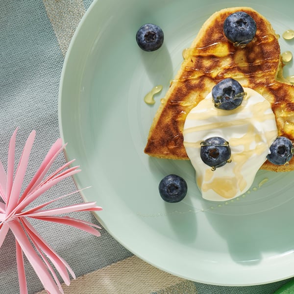 Light green kids plate holds delicious blueberry pancake topped with yoghurt and drizzled with honey, all placed on a cosy blue tablecloth.