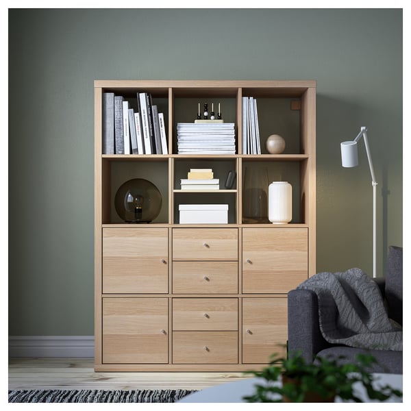 KALLAX light wooden bookshelf with shelves and drawers, standing against green wall, surrounded by larger grey chair and small plant.