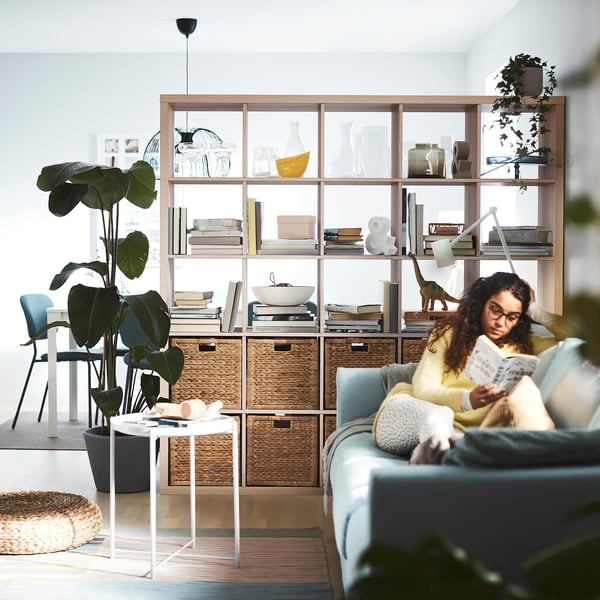 Person reading on blue couch near wooden shelf with baskets and decor, green plant, and white side table.