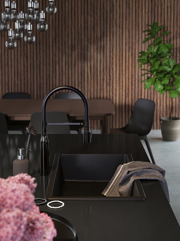 Modern kitchen sink, matte black quartz, wooden backsplash, black chairs, green plant.