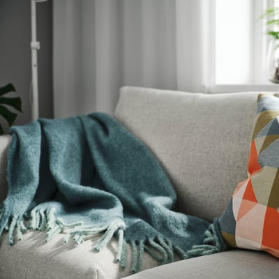 Grey-turquoise throw blanket on light couch, with soft, geometric pillow. Warm, cosy atmosphere.