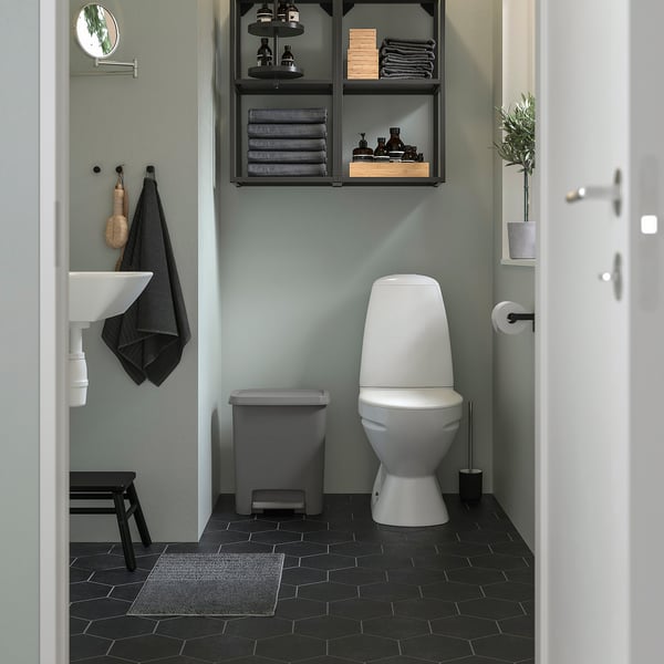 Black bathroom with white toilet, grey bin, green walls, black floor tiles, towels on chair and shelf.