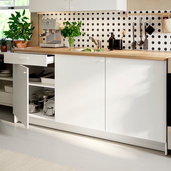 Modern kitchen with white cabinets, oak countertop, and polka dot backsplash. Pots, tools hang, plants decorate. Open cabinet reveals pots and pans.