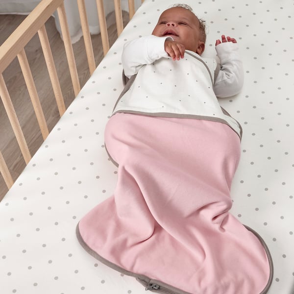 Baby in soft, pink sleep bag, lying on a crib with light dot patterns; designed for cosy sleep.
