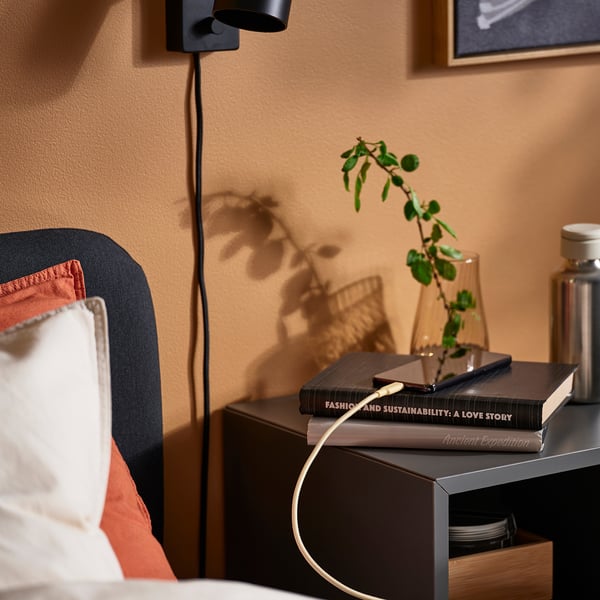 Cosy bedroom scene: a side table with a small potted plant, a stack of books, and a black wall-mounted lamp plugged into a long usb-c cable.