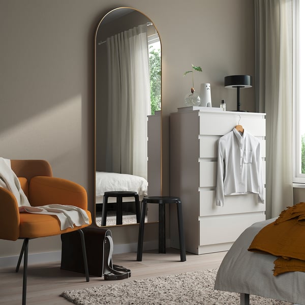 LINDBYN gold-framed mirror leaning on wall, beside white dresser, orange chair, and rug.