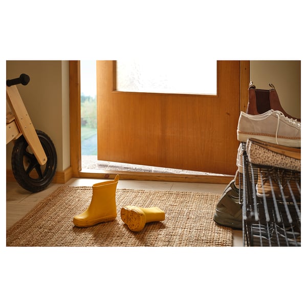 A neutral-toned jute rug with yellow rain boots on it, next to a shoe rack and a wooden walker.