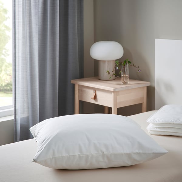Bedroom with large white LUDDROS pillow protector on bed, light wood bedside table, modern lamp & plant. Curtains and grey wall in background.