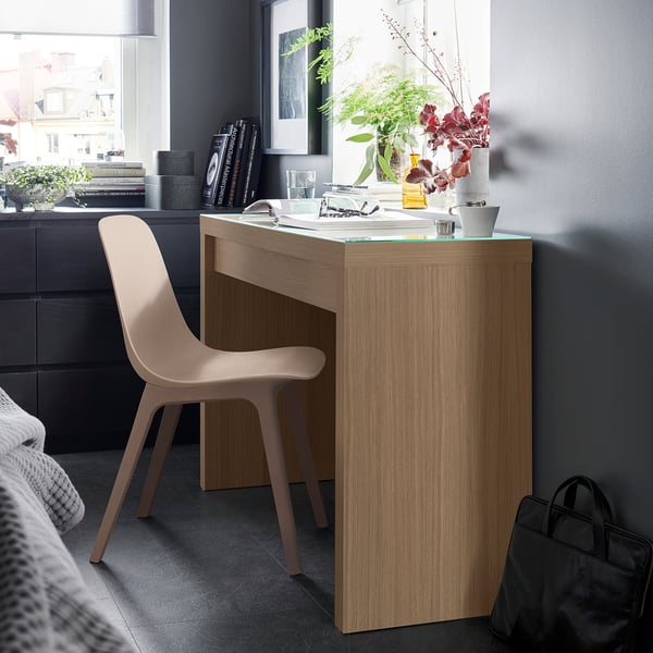 Modern desk in natural wood tone, narrow size, light-colored chair, flowers, books, and city view.