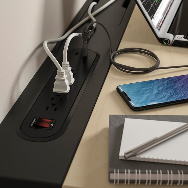 A modern desk with built-in power outlets and a cable management tray, showcasing a smartphone, notebook, and a computer monitor.