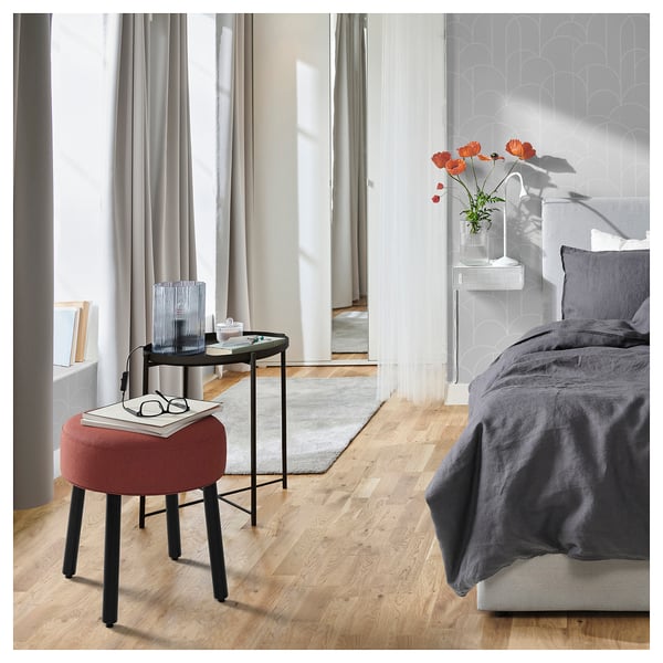 Modern bedroom with MÖRTFORS rusty-red stool, grey bed, side table, and floral vase. Light wood floor.