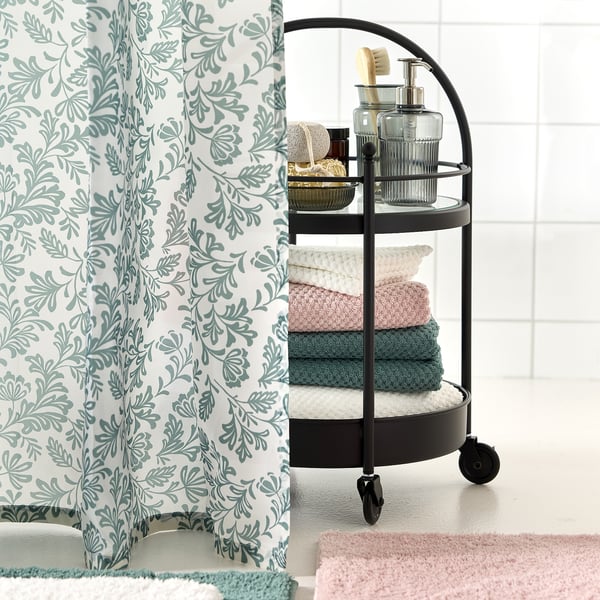 Elegant bathroom with leafy green shower curtain. Black cart stores towels and toiletries, adding contrast to light tiles.