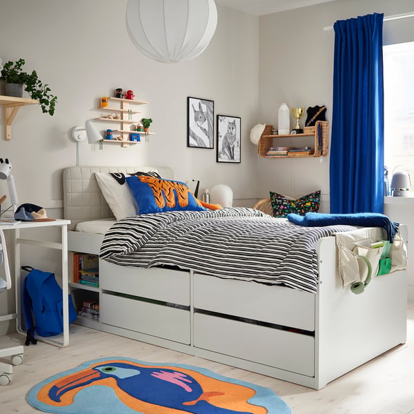 Kids room with white bed, blue curtains, colourful pillows, wooden floor, desk, plants, cosy.