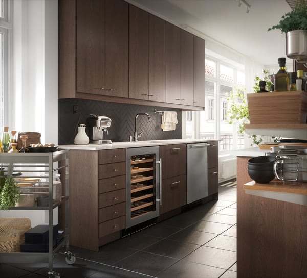 Modern kitchen with wooden cabinets, stainless steel appliances, herringbone backsplash, and a cart with plants and cooking supplies.