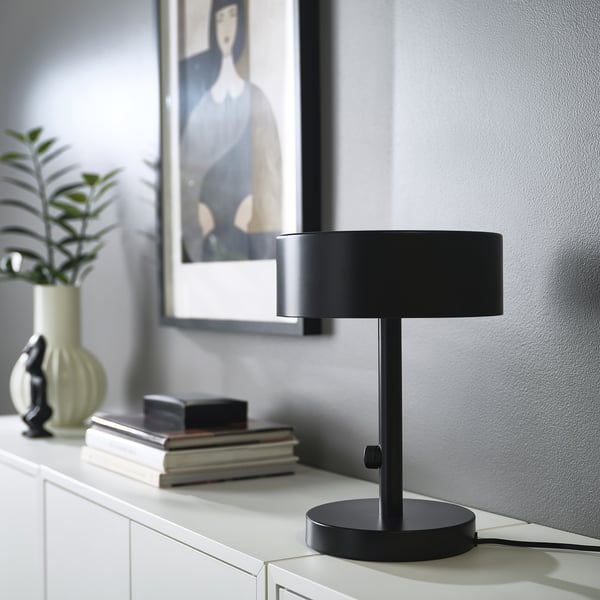 Black NYMÅNE lamp sits on shelves with books & vase, smaller than books & plant.