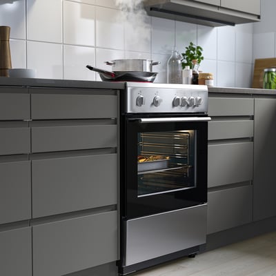 Stainless steel ÖRMINGE oven in modern kitchen, sits between grey cabinets, with food inside and pots on top.
