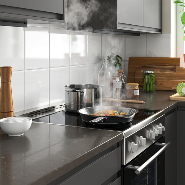 Modern kitchen with black cooktop, hood, and silver knobs. Dark counter with wok pan, food, and greenery.