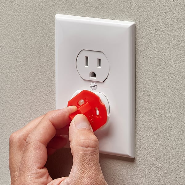 A hand instals a red child safety plug into a wall outlet to prevent access.
