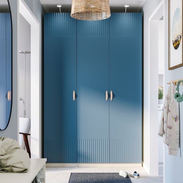 Modern room with tall blue PAX wardrobe, gold handles, shoe rack, chair with clothes, wicker pendant light, and round mirror.