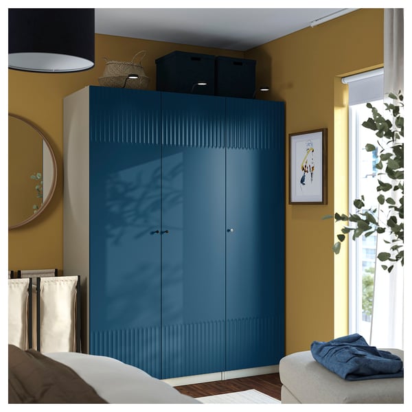 Blue PAX wardrobe near ceiling, sleek design, mirror left, plant right, bed with bedding, towel, picture on wall, natural light.