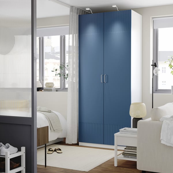 Modern bedroom with PAX wardrobe in deep blue. Tall, sleek storage unit fits between windows, complementing the minimalistic decor.
