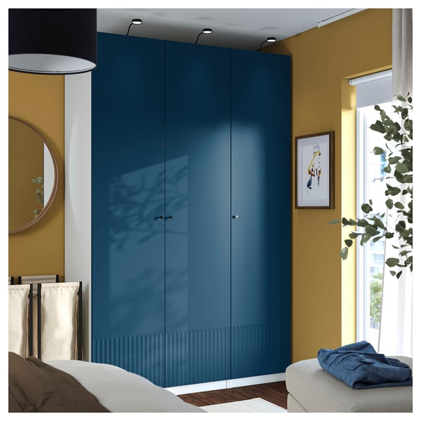 Bedroom with tall blue wardrobe, white bed, plant, mirror, and window.