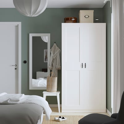 White PAX wardrobe in bedroom. Tall, slim, matching white room decor. Contains mirror & coat rack. Placed against sage green wall.
