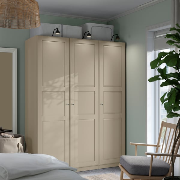 Beige IKEA PAX wardrobe in green bedroom, larger than nearby rocking chair, three doors, mesh baskets, window with wooden chair.