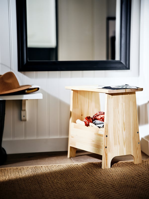 A light wooden stool named PERJOHAN, with simple design and cut-out handle, placed in a room with a mirror and hat on a shelf.