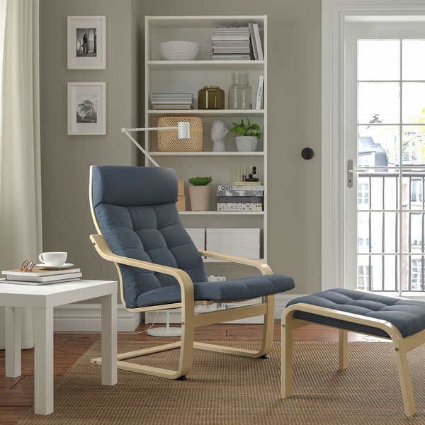 Modern living room with grey POÄNG armchair, matching footstool, white side table, and bookshelf. Large window with natural light.