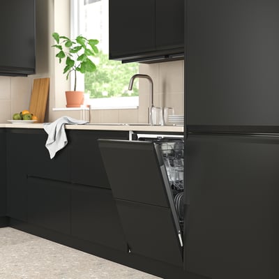 Modern black kitchen with dark cabinets, open RANSBERG dishwasher, white countertop, potted plant, fresh fruits, natural light.
