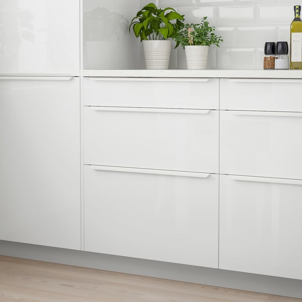 Modern white kitchen with sleek, glossy drawers against wooden floor. Small plants and bottle decorate countertop.