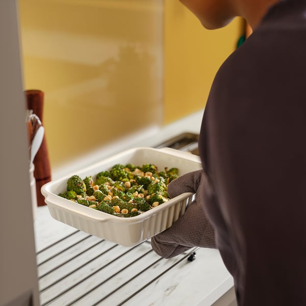 Person holding oven dish with broccoli, using black oven glove, near kitchen stove.