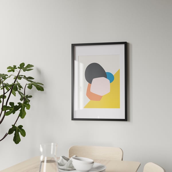Framed abstract art hangs on a wall. Wooden dining table with chairs and dishes below it. Green leafy plant on the left side.