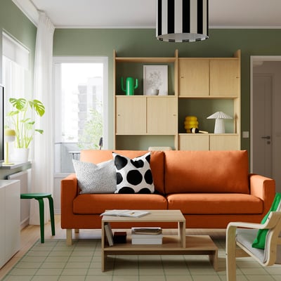 Modern living room with large orange SALTSJÖBADEN sofa, light wood coffee table, and storage shelves. Bright, airy decor with green accents.