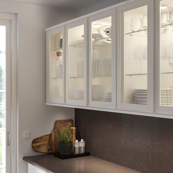 Modern white glass-door kitchen cabinet with metal frame, containing dishes. Below, dark countertop holds wooden boards, plant, and bottles.