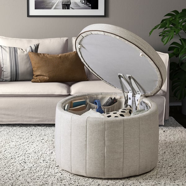 White upholstered storage ottoman open, revealing tools inside, next to a beige sofa with pillows on a light carpeted floor.