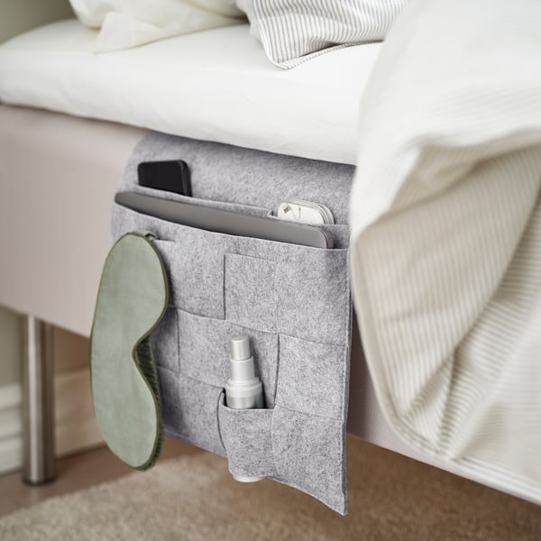 Grey, felt bed organiser with multiple pockets holding a tablet, phone, eye mask, and spray bottle, designed to fit under mattresses.