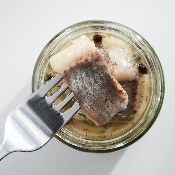 Small jar with marinated herring, preserved in dill and onions. Silver fork inserted, lighter grey fish, light yellow liquid.