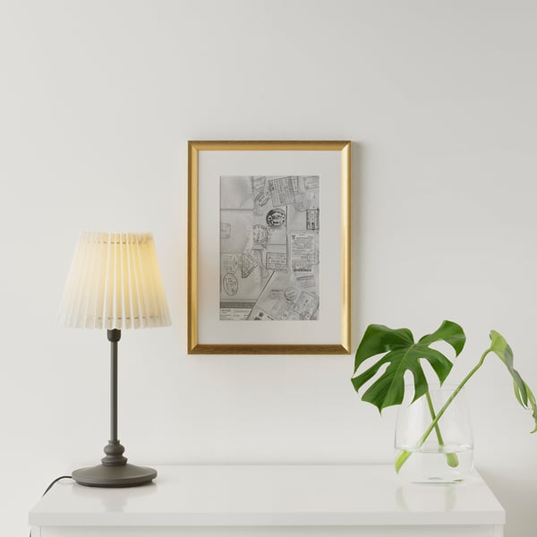 Framed poster on white wall with gold frame. Table below has black lamp with beige shade and green plant in clear vase.
