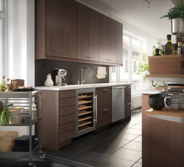 Modern kitchen with dark wood SINARP cabinets, stainless appliances, herringbone tile backsplash, natural light, and plants.