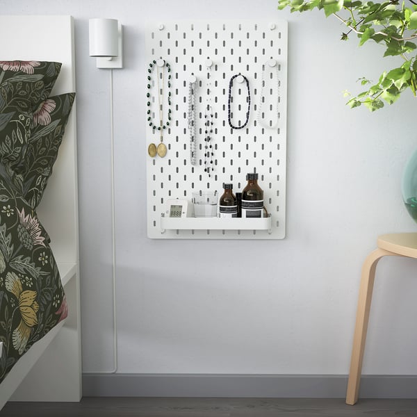 SKÅDIS pegboard with jewellery, bottles, clock. Beside bed with floral pillows and chair.