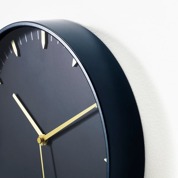 Modern wall clock with gold hands on dark face, mounted on white wall.