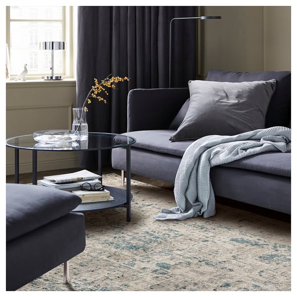 Modern living room with grey SKAFTERUP rug, grey sofa, glass coffee table, and decorative vase.