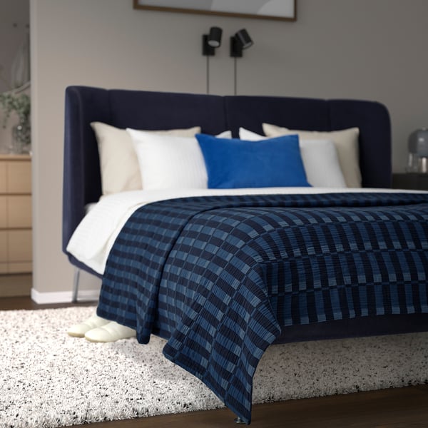 Bed with dark blue chequered SKOGSKOVALL blanket, white pillows, and large dark headboard. White rug lies underneath, adding warmth to the room.