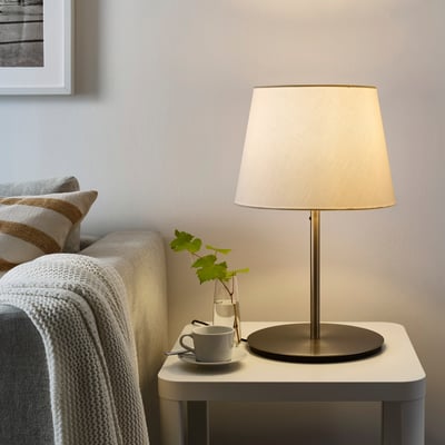 A SKOTTORP lamp with beige shade on side table, surrounded by cup, vine leaves, and cosy couch with white blanket. Soft glowing plastic shade.