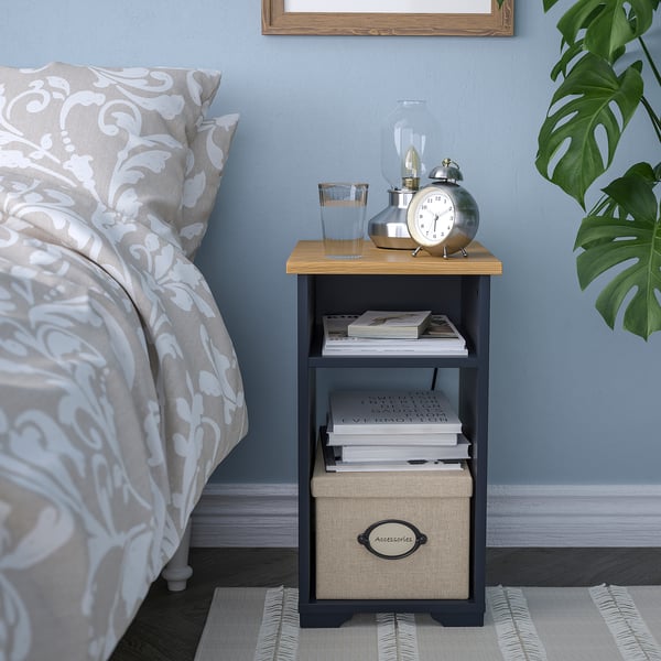 SKRUVBY side table with wooden top, open shelves & woven box, beside bed holding books, clock, lamp.